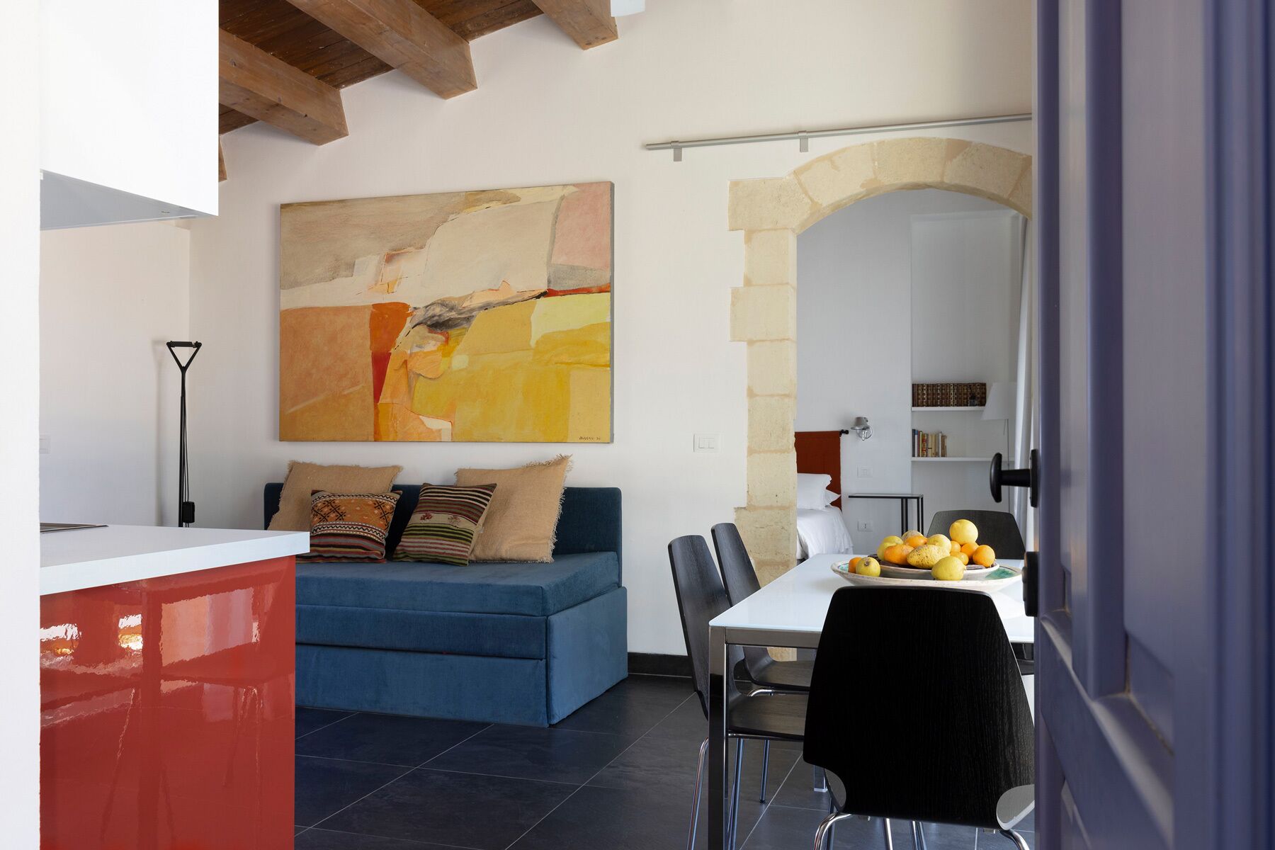 A modern living room with a blue sofa, a dining table with chairs, and a large abstract painting on the wall.