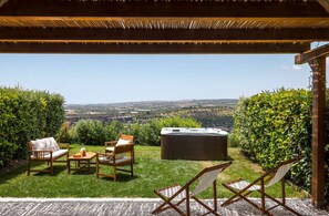 A patio with a hot tub, wooden chairs, and a table with drinks, overlooking a scenic landscape.