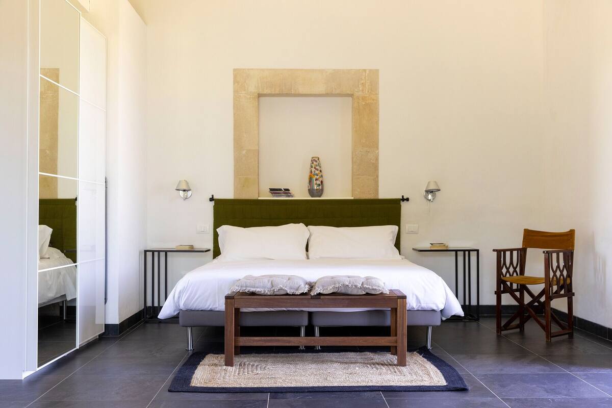 Quarto moderno com uma cama grande, um banquinho, uma cadeira e um espelho.