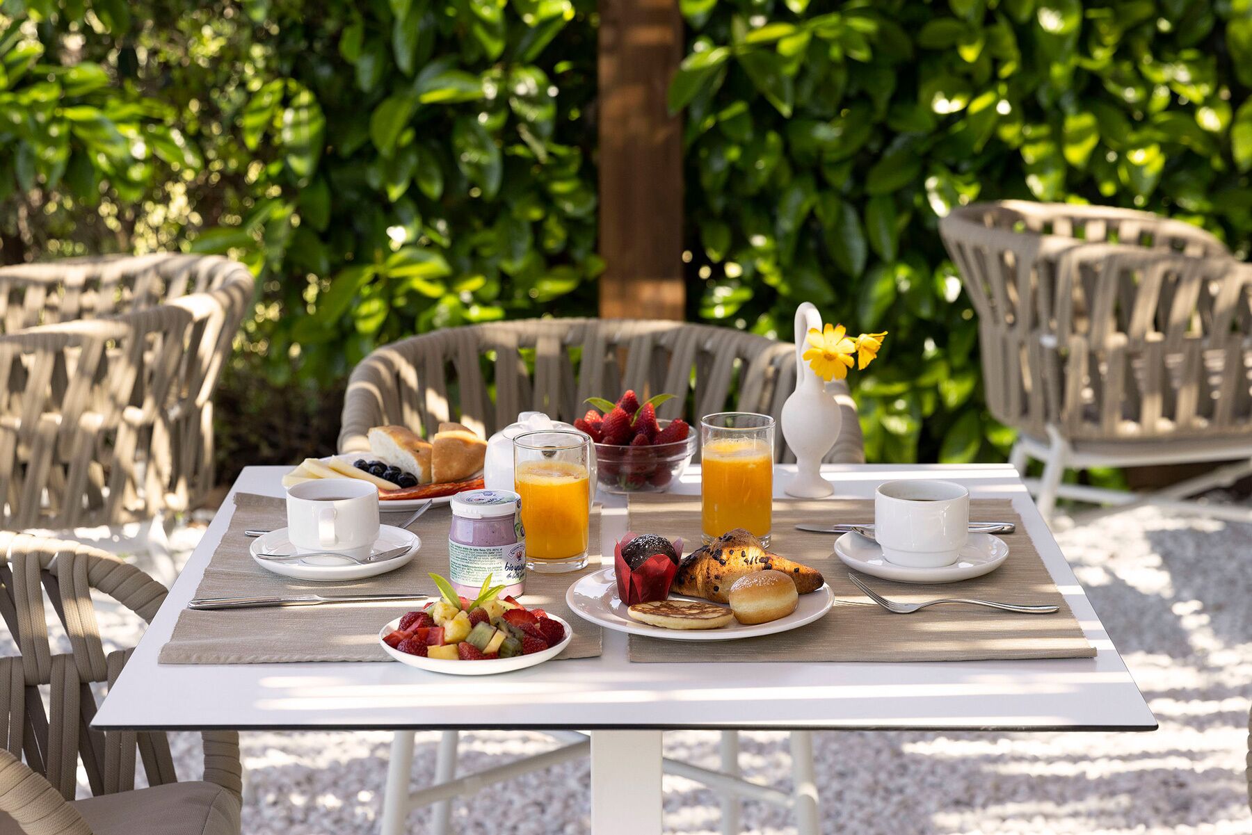 An outdoor breakfast setting with a table set for two, including plates of fruit, pastries, and beverages.