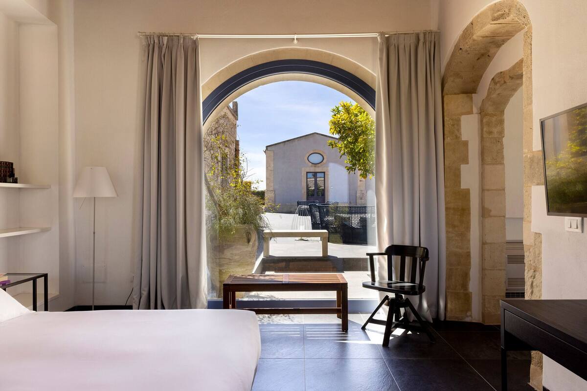 Quarto de hotel com uma cama grande, uma escrivaninha, uma cadeira e vista através de uma porta arqueada para uma área externa com um edifício e vegetação.