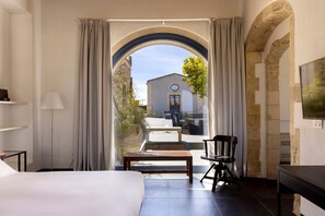 A hotel room with a large bed, a desk, a chair, and a view through an arched doorway to an outdoor area with a building and greenery.