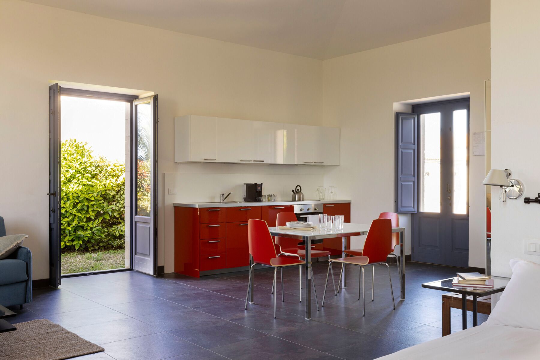 A compact kitchen with red cabinets and a white countertop, a dining area with red chairs, and a large window providing natural light.