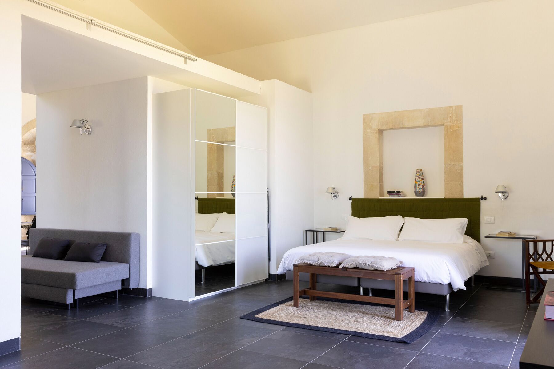 A modern bedroom with a bed, a small wooden bench, a grey sofa, and a large mirror.