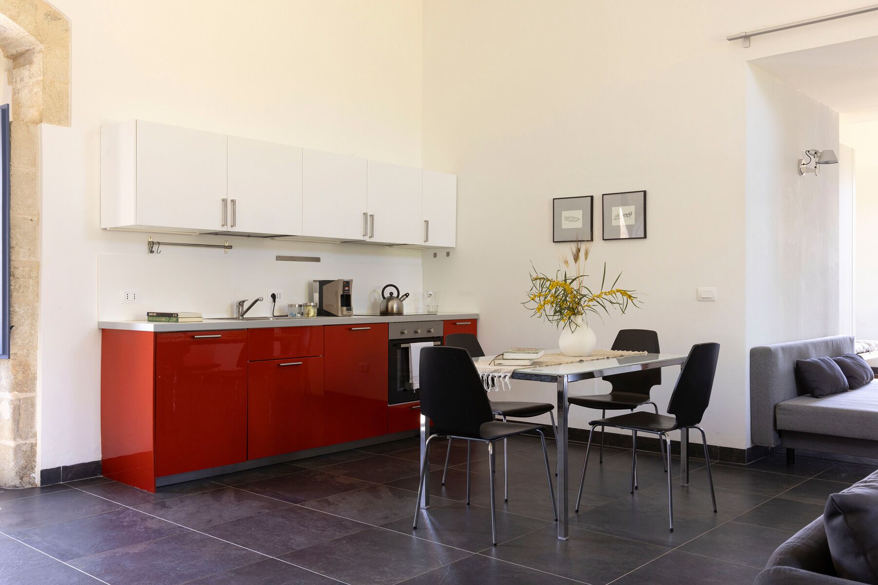 A modern kitchen with white and red cabinets, a dining table with four chairs, and a grey sofa in a living area.