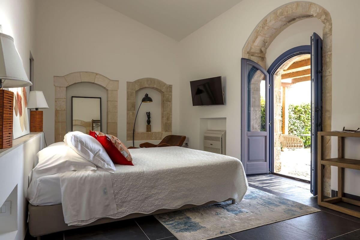 Quarto com cama grande, televisão e vista para área externa por uma porta aberta.