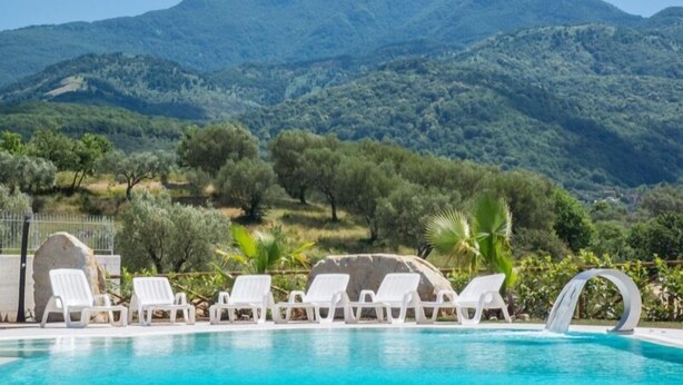 A pool area with white lounge chairs and a waterfall feature, surrounded by lush greenery and mountains in the background. - Agriturismo L'Oasi (Ceraso)