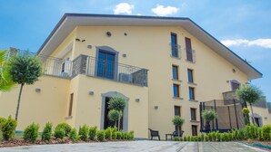 A two-story building with a flat roof, multiple balconies, and a paved courtyard with small trees and shrubs. - Agriturismo L'Oasi (Ceraso)