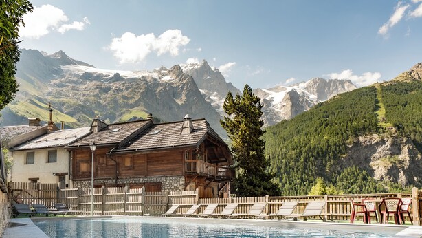 A pool area with lounge chairs, a wooden building, and a mountainous backdrop. - Hôtel Castillan (La Grave)