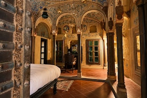 Quarto com cama, uma cadeira e escrivaninha com abajur. Apresenta colunas decoradas e piso de azulejos.