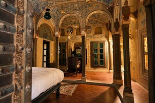 Una habitación con una cama, una silla y un escritorio con una lámpara. Se observan columnas decorativas y un piso de baldosas.