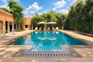 Una piscina rectangular con agua azul cristalina, rodeada de sillones de descanso y sombrillas, con un pavimento de baldosas y una pared de piedra en un lateral.