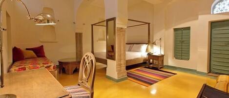 A spacious room with a large bed, a desk, and a chair. There is a window with green shutters and a ceiling fan.