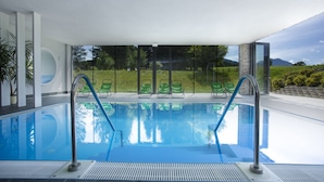 Una piscina interior con paredes de vidrio transparente, sillas de descanso verdes y vistas a una zona ajardinada con árboles y montañas al fondo.