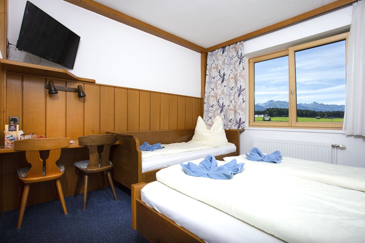 Quarto de hotel com duas camas, uma escrivaninha com cadeiras, televisão e janela com vista para as montanhas.