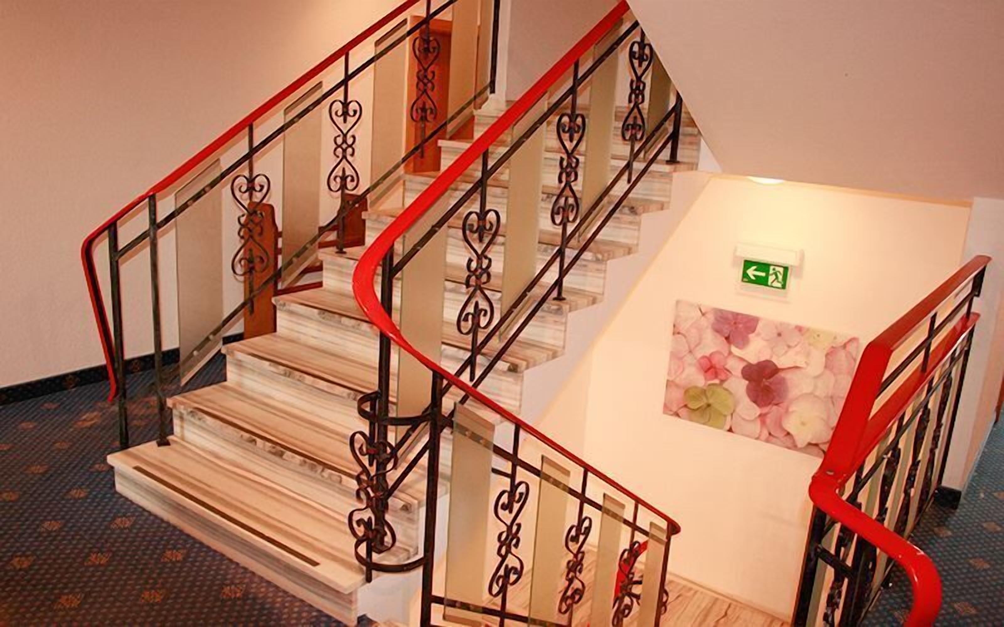 A staircase with ornate metal railings and a red accent, leading to a landing with a floral artwork and an emergency exit sign.