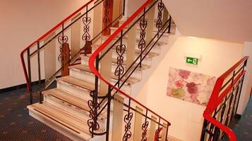 Una escalera con barandillas de metal decoradas y un detalle rojo, que conduce a un rellano con una obra de arte floral y un letrero de salida de emergencia.