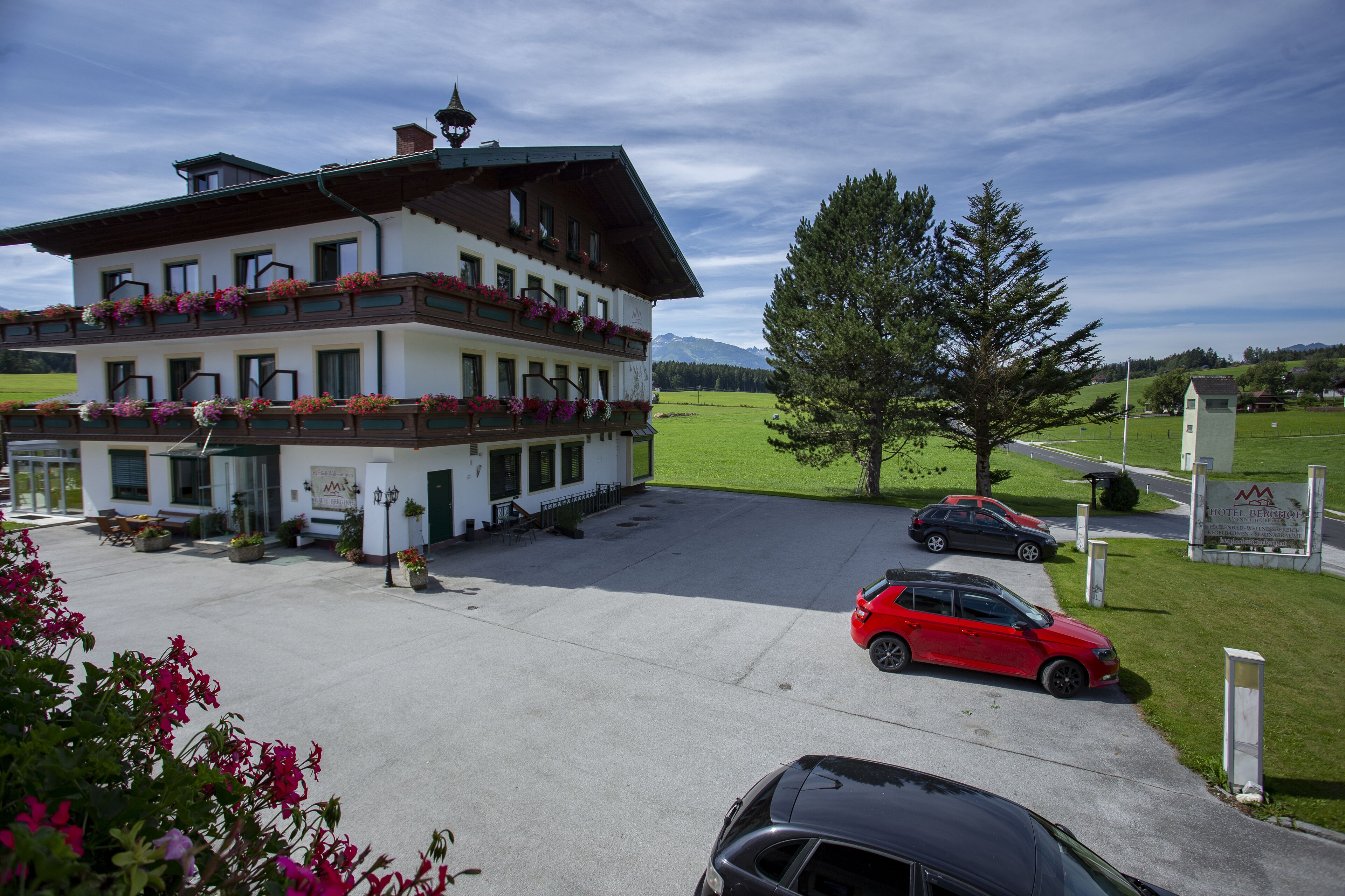 Un hotel de varios pisos con un balcón decorado con flores, un estacionamiento con coches y un cartel que indica el nombre del hotel.