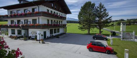 A multi-story hotel with a balcony adorned with flowers, a parking area with cars, and a sign indicating the hotel's name.