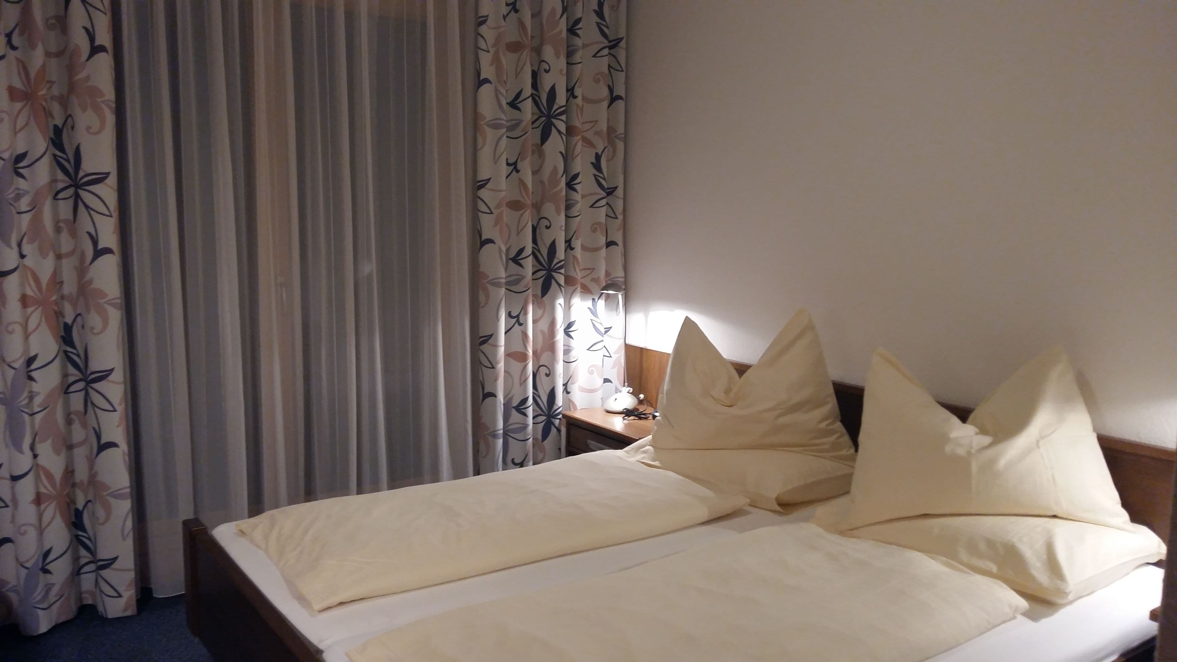 Ein Doppelbett mit zwei Kissen, ein Nachttisch mit Lampe und blumige Vorhänge.