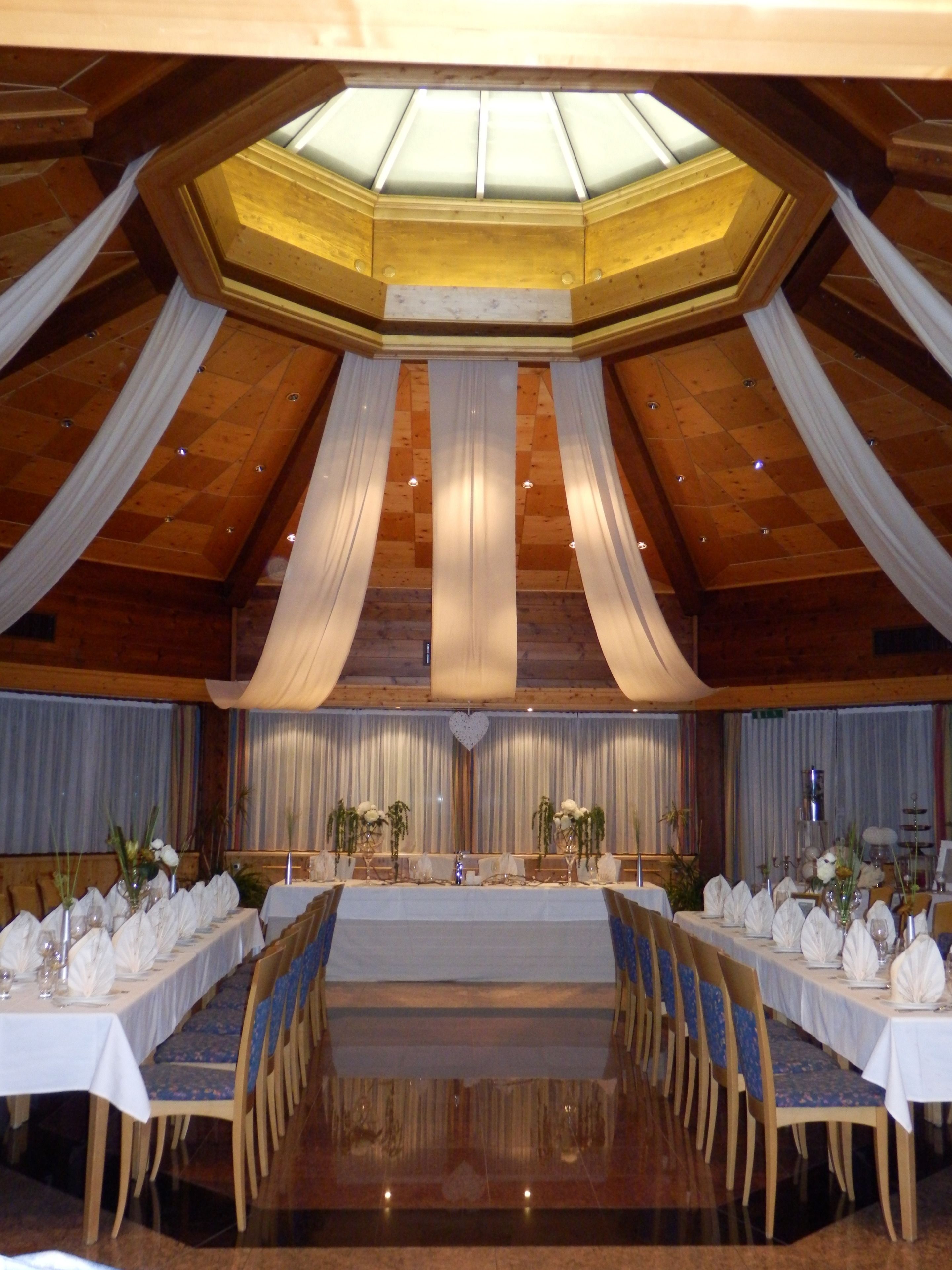 A banquet hall with a high ceiling, wooden beams, and a central stage. Tables are set with white tablecloths and chairs.