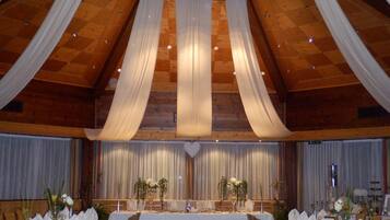 A banquet hall with a high ceiling, wooden beams, and a central stage. Tables are set with white tablecloths and chairs.