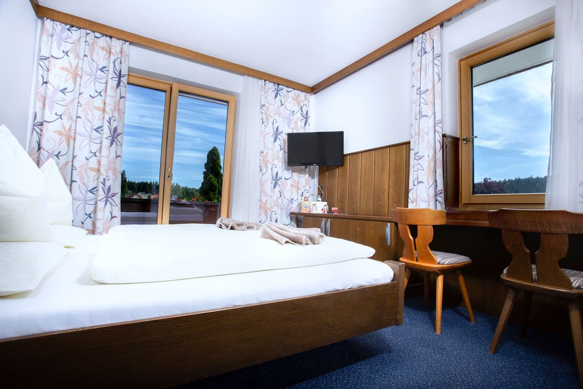 Quarto de hotel com cama, escrivaninha, cadeira, televisão e amplas janelas.