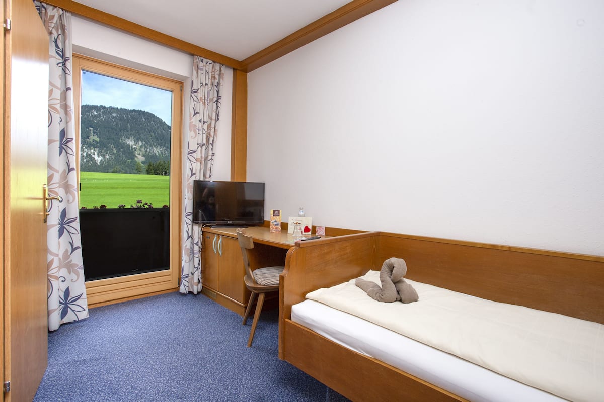 Quarto com cama, escrivaninha, cadeira, televisão e janela com cortinas.