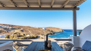 Une table en bois ornée d’une assiette décorative et d’un vase, des chaises en osier et une vue sur la mer et un paysage vallonné.
