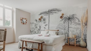 Quarto com cama, um banquinho de madeira, uma mesinha e um tapeçário na parede.