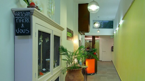 A hallway with a sign reading "ARTISAN NACRE & VOUS", a large potted plant, and a reception area with a person behind the counter. - Hotel Palym (Biarritz)
