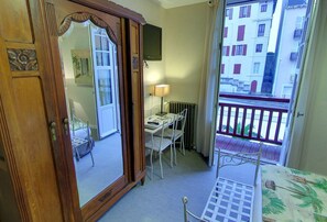 A room with a wooden wardrobe, a bed, a desk with a chair, a table, and a balcony with a view of buildings. - Hotel Palym (Biarritz)