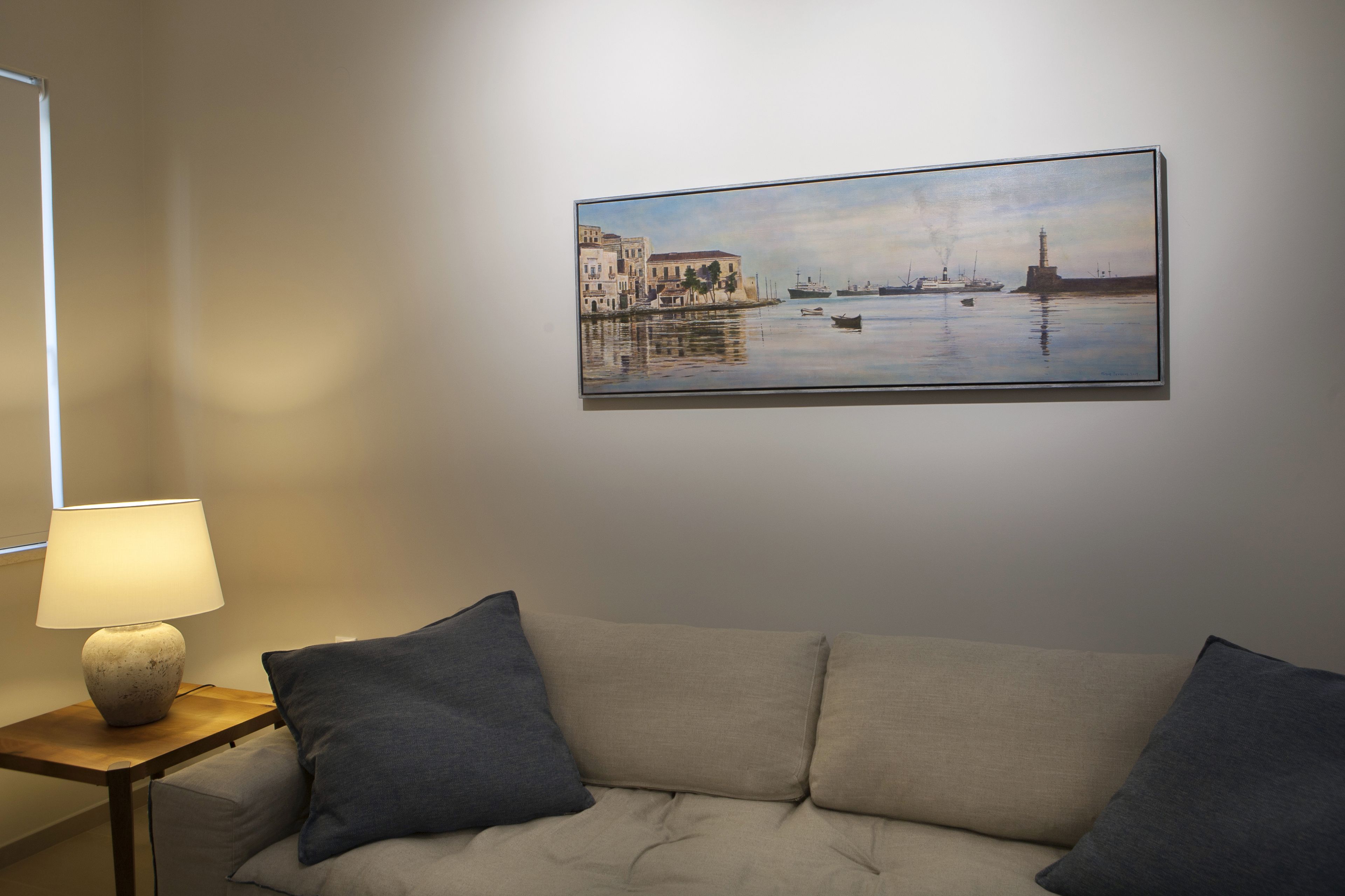 Ein Zimmer mit einem beigen Sofa, einem kleinen Tisch mit einer Lampe und einem Gemälde einer Hafenszene an der Wand.