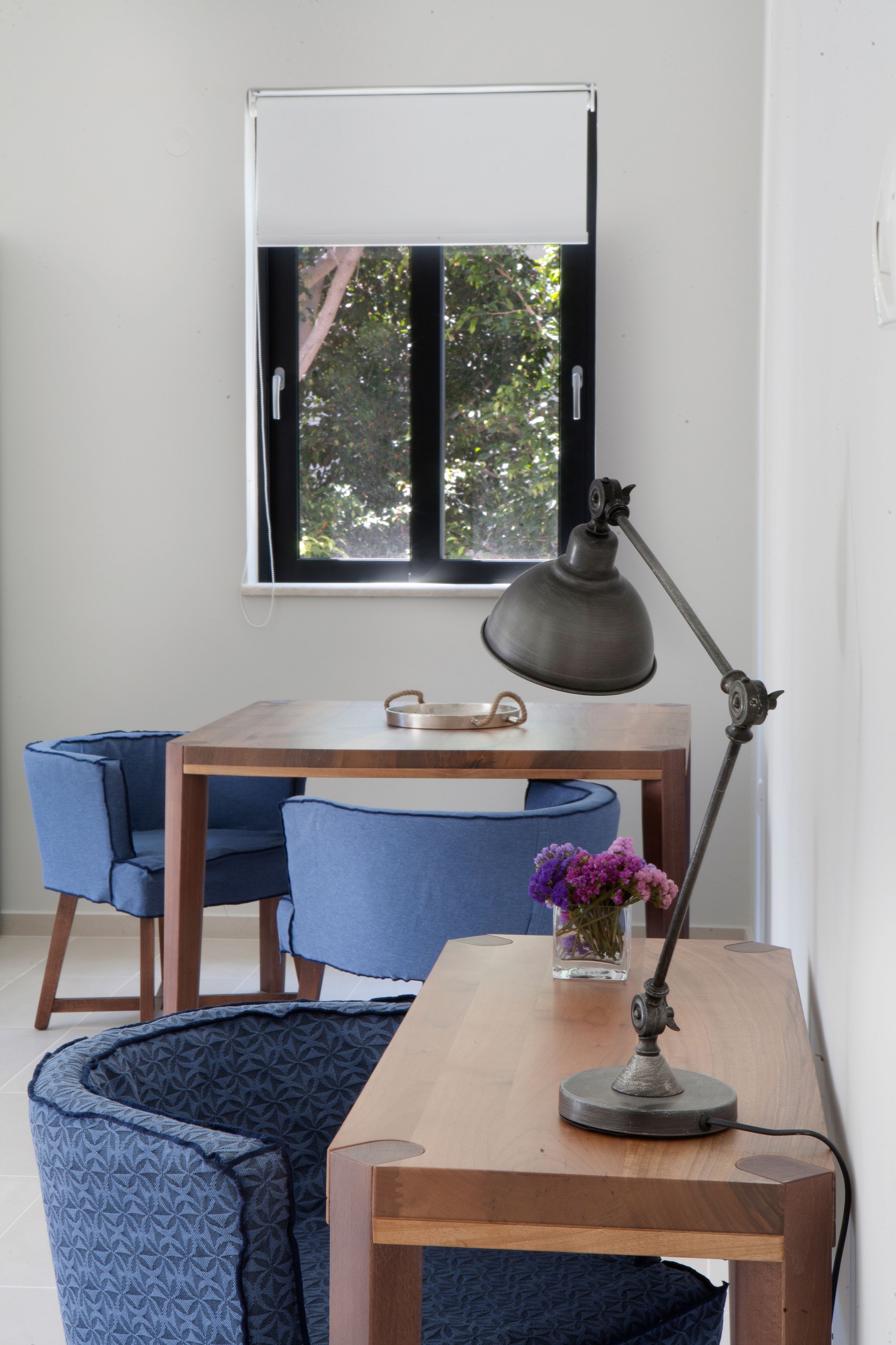 A room with a wooden desk, a blue chair, a blue couch, a window with a view of trees, and a desk lamp.