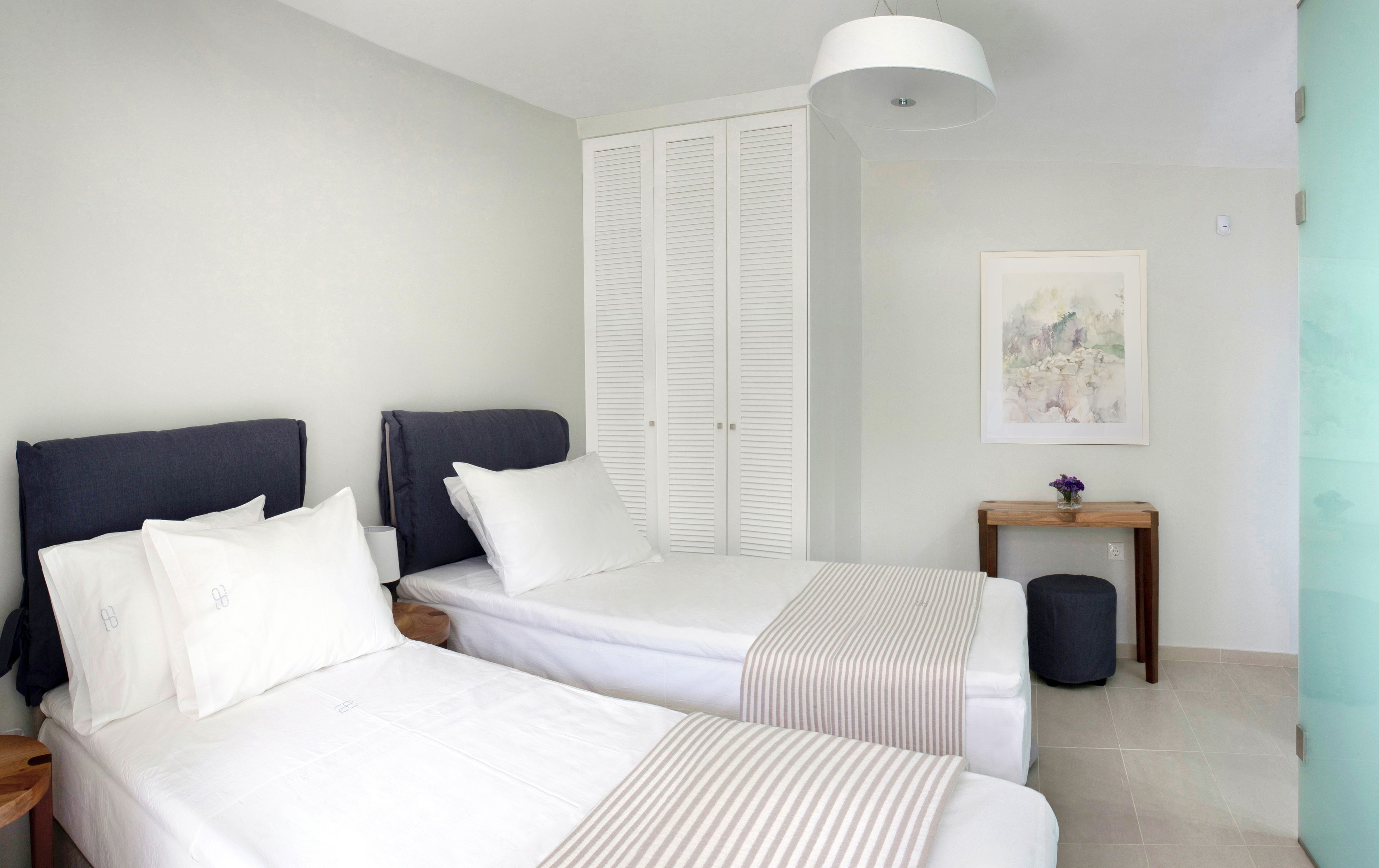 A bedroom with two beds, a bedside table, a lamp, a picture on the wall, and a small wooden table with a vase.