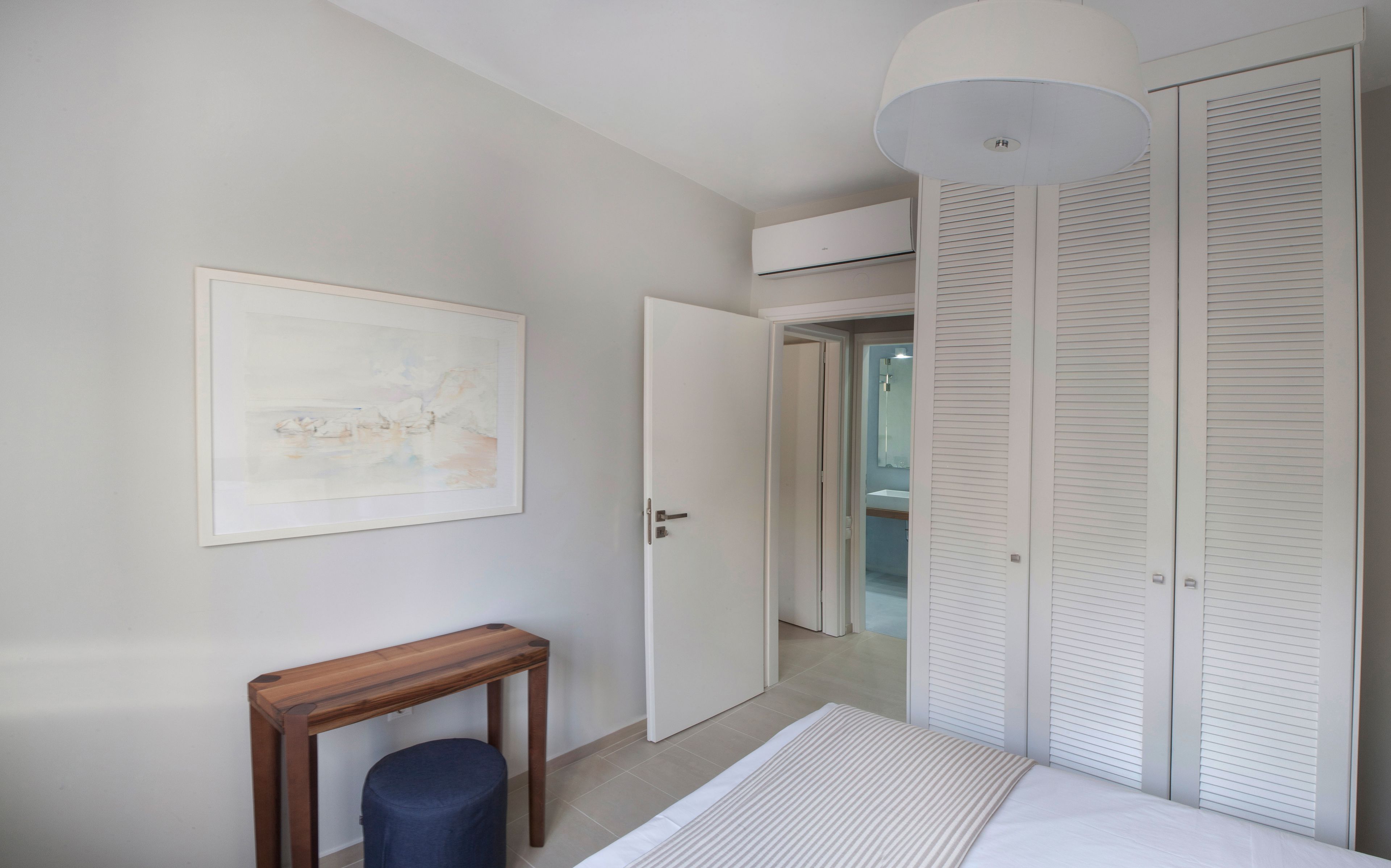 A bedroom with a bed, a wooden side table, a blue ottoman, a white ceiling light, and a white air conditioning unit.