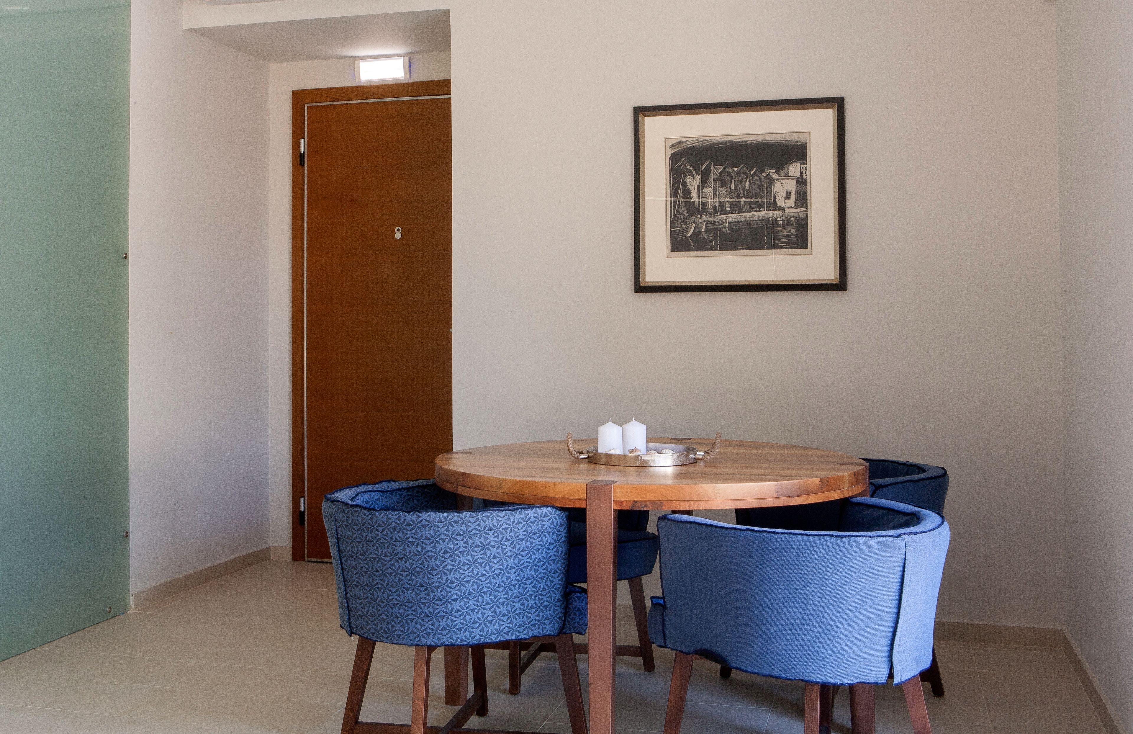 A dining area with a round wooden table, four blue upholstered chairs, a framed black and white artwork on the wall, and a wooden door in the background.