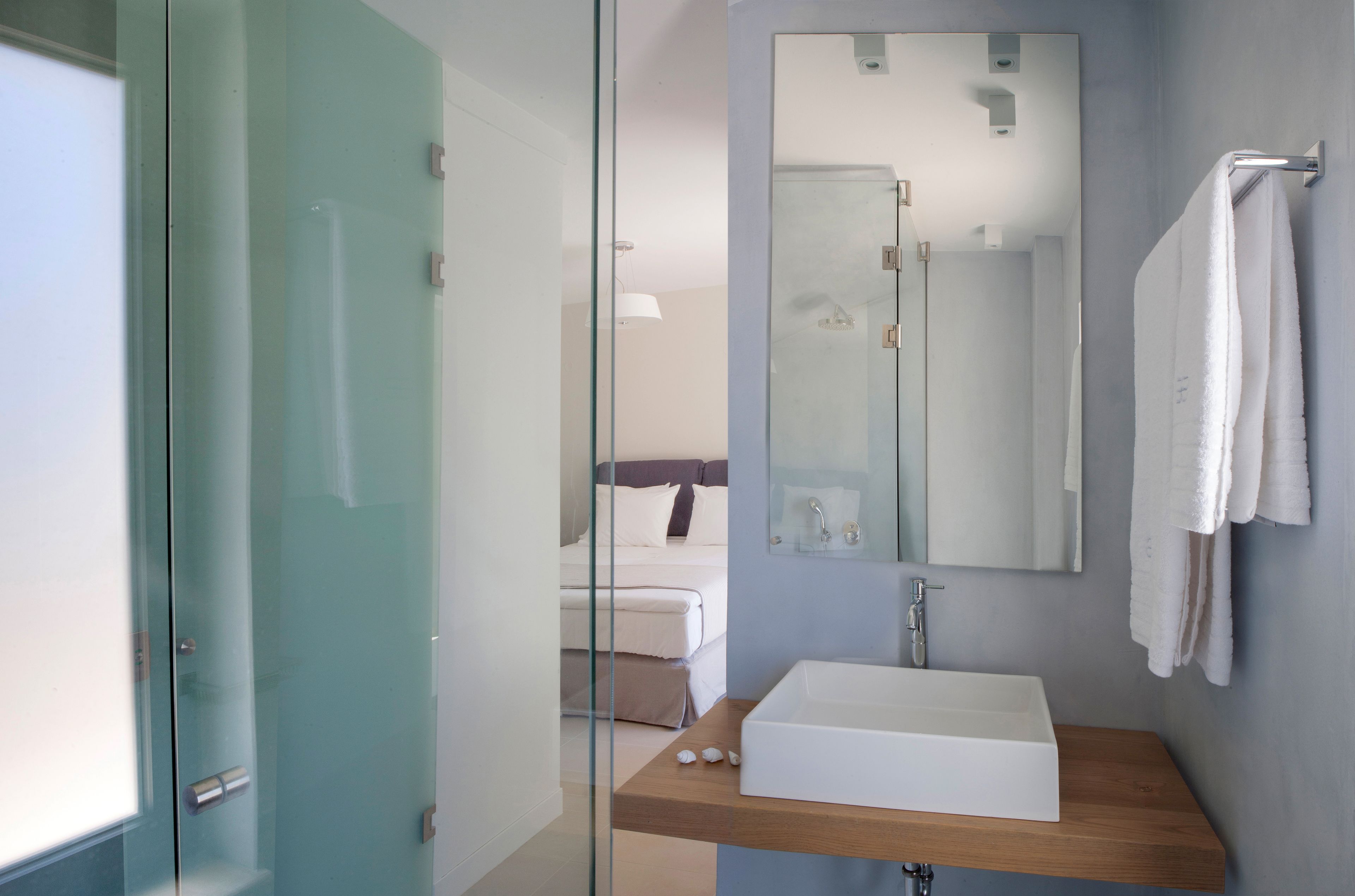 A modern bathroom with a glass shower enclosure, a wooden vanity, a large mirror, and a bed in the background.