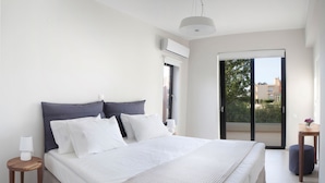 A modern bedroom with a large bed, a bedside table with a lamp, and a view of the outdoors through a sliding door. - Aethrion Homes (Chania)