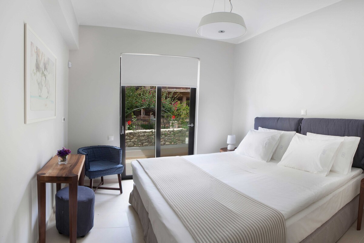 Quarto com cama, escrivaninha, cadeira, luminária e vista para um jardim através de uma porta deslizante.