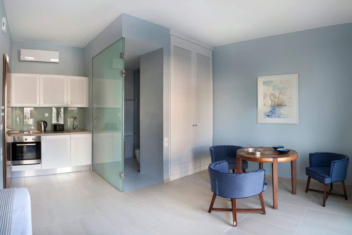 Una cucina moderna con mobili bianchi, forno integrato e una porta vetrata che dà su un bagno. È presente un'area pranzo con un tavolo rotondo in legno e due sedie blu.