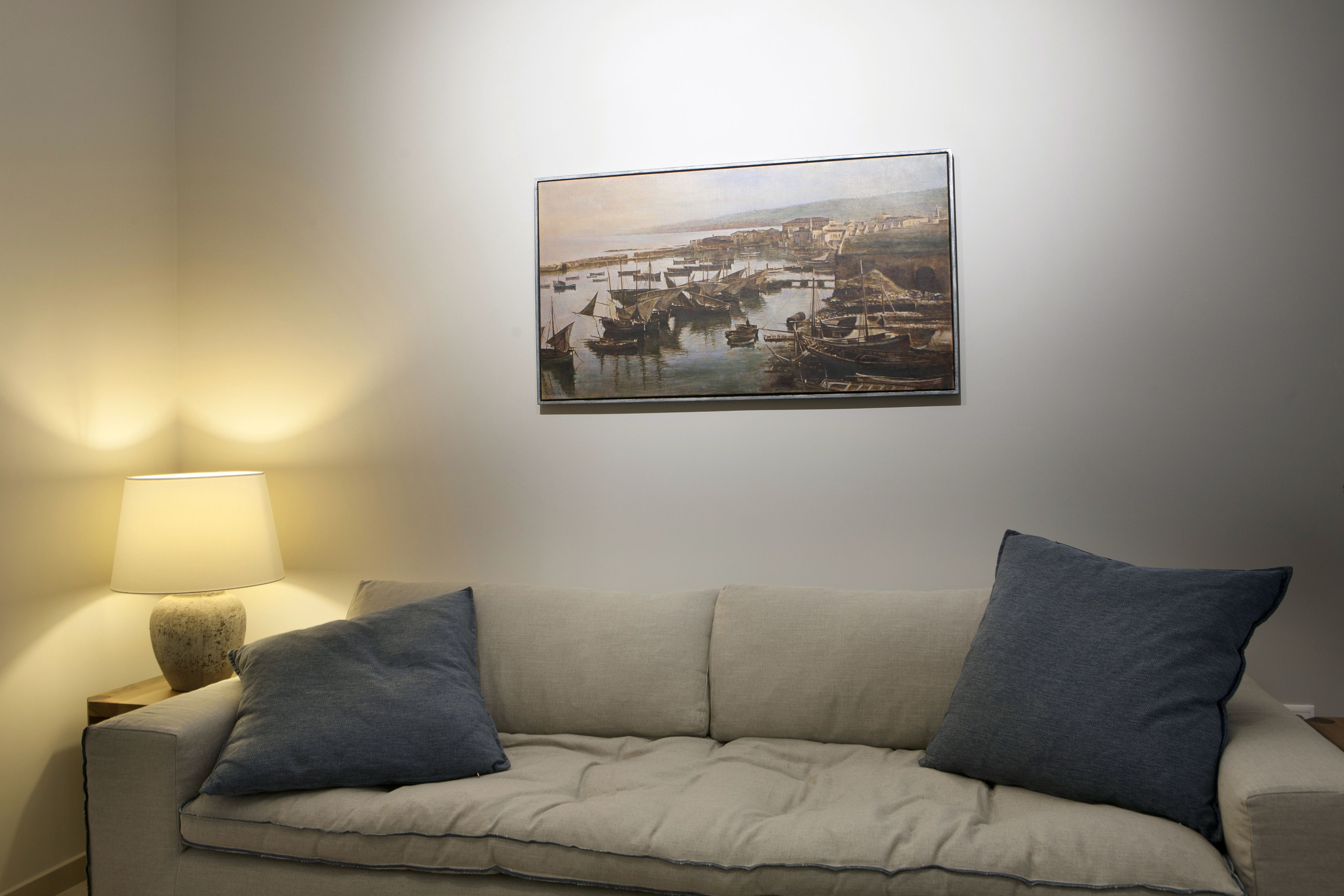 A living room with a beige sofa, two pillows, a lamp, and a framed painting of a harbor scene.