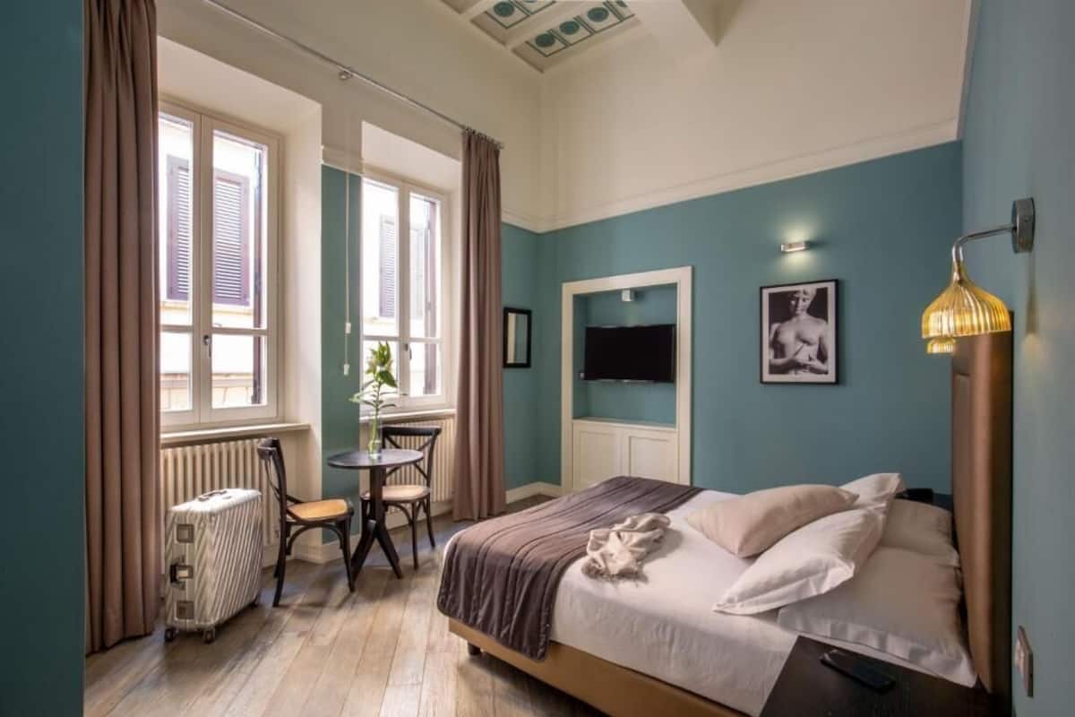 Un dormitorio con una cama, un escritorio y una silla. Hay una ventana con cortinas, un radiador y un televisor montado en la pared.