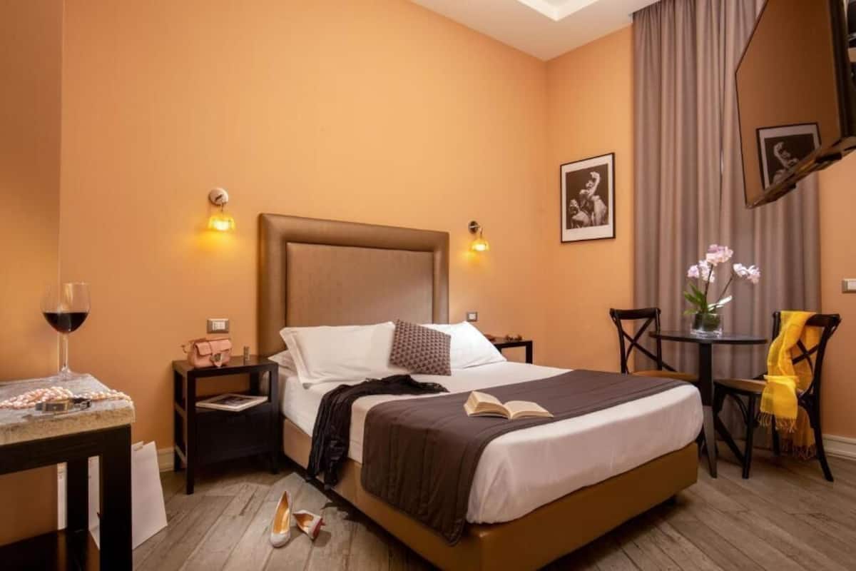 Una habitación de hotel con cama, mesitas de noche, una pequeña mesa con una copa de vino, una silla y un escritorio.