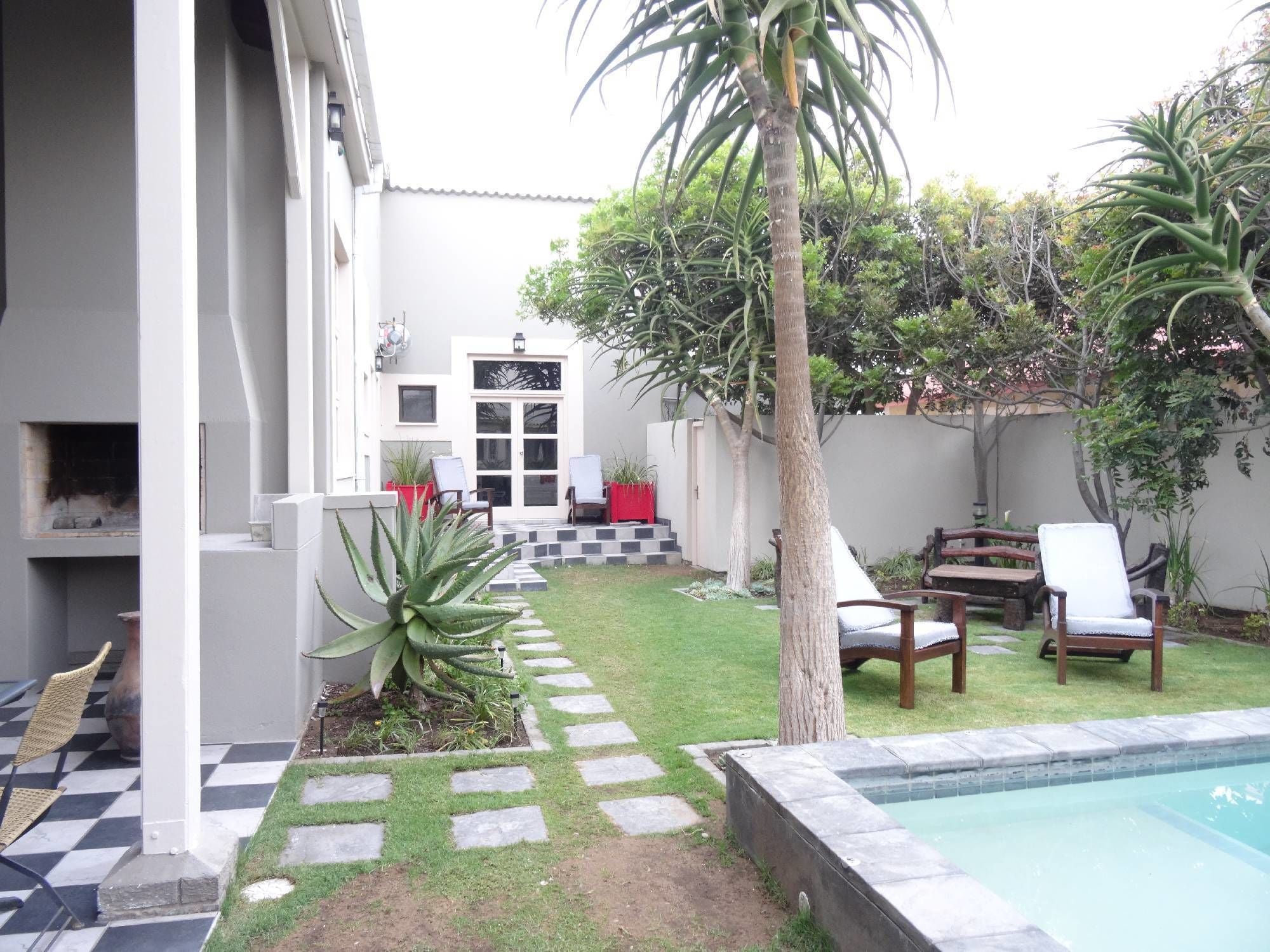 Ein Garten mit Swimmingpool, einer Terrasse mit Stühlen, einem kleinen Garten und einer Tür zu einem überdachten Bereich.