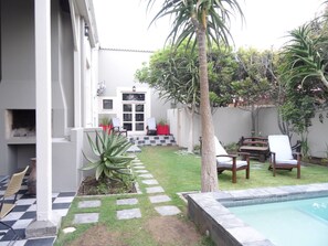 A backyard with a swimming pool, a patio with chairs, a small garden, and a door leading to a covered area.