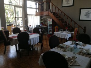 A dining area with multiple tables set for meals, a staircase, and a refrigerator with beverages.