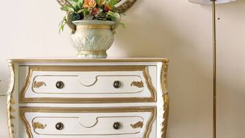 A white dresser with gold trim and two drawers, a vase with flowers, a mirror, and a floor lamp.