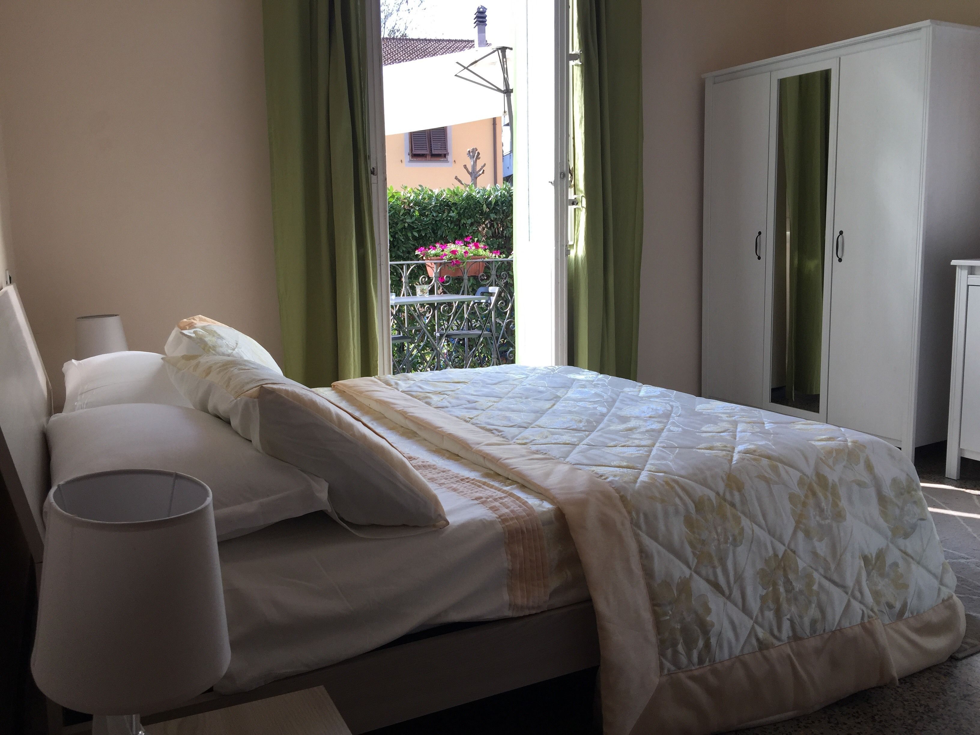 Una cama bien hecha con edredón y almohadas blancos, una lámpara de noche y vistas a un balcón con cortinas verdes y barandilla.