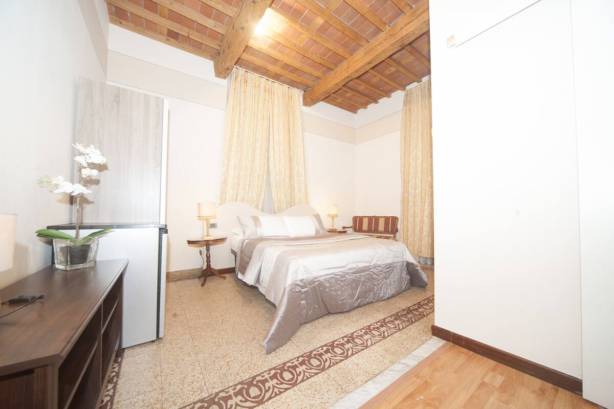 Une chambre à coucher avec un lit, des tables de chevet, un bureau, une plante en pot et une fenêtre avec des rideaux.